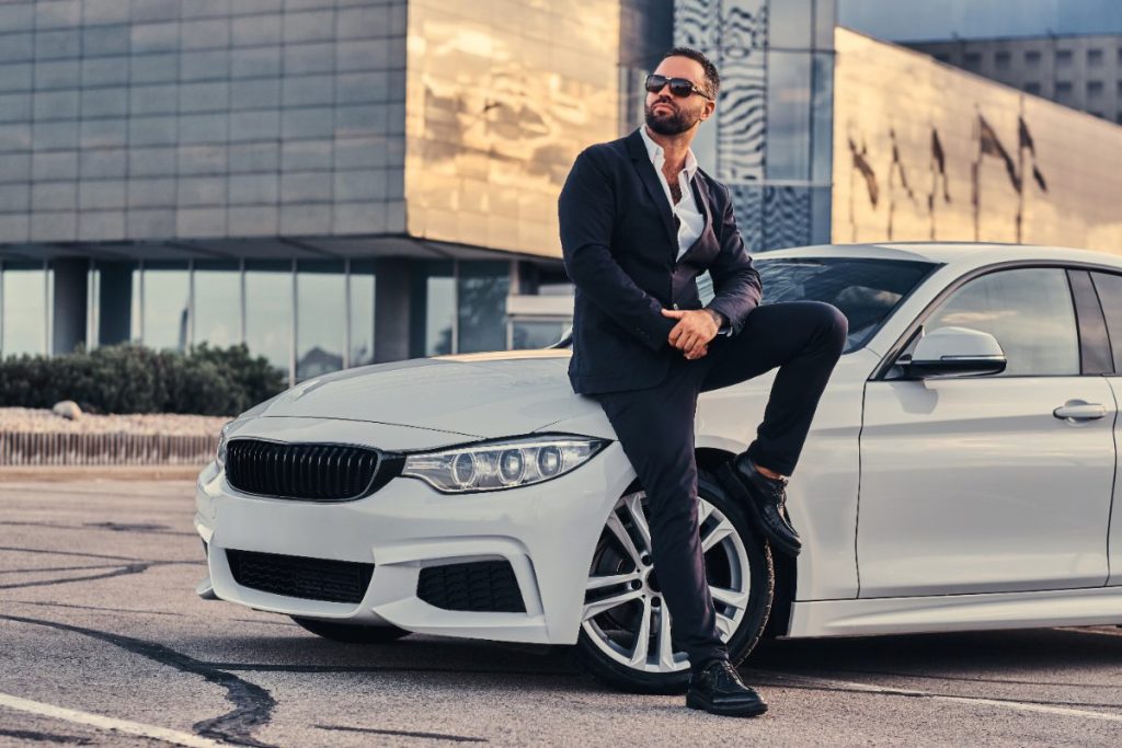 Un homme barbu appuyé contre une voiture de luxe